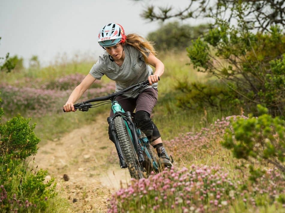female mountain biker riding through dirt trail with bushes and flowers